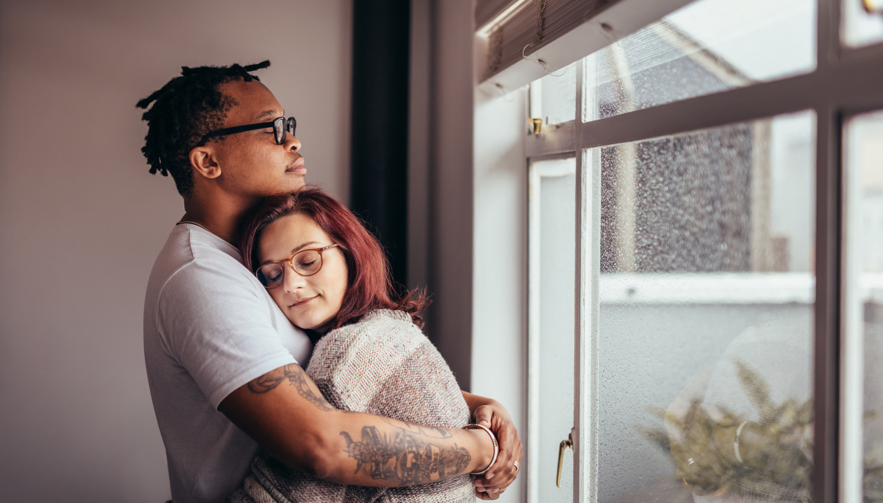 Interracial couple hugging each other while standing near window. African man embracing his caucasian girlfriend at home.